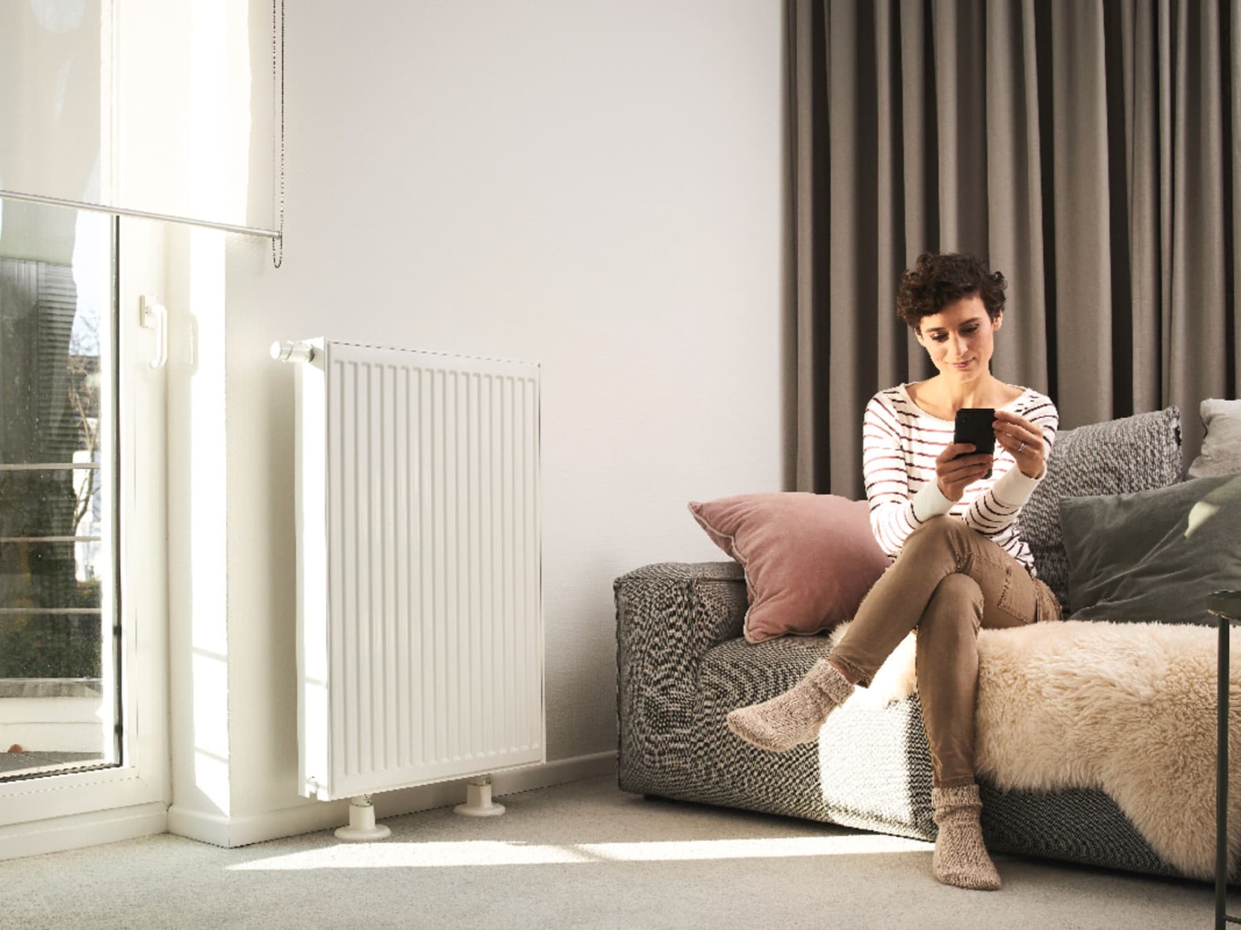 Kobieta siedzi na sofie i korzysta ze smartfona. Sofa znajduje się przed oknem i obok grzejnika.